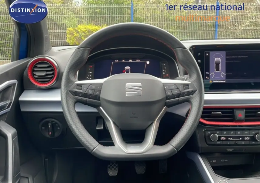 Vue centrée du volant perforé et tableau de bord numérique d’un SEAT Arona bleu saphir 2025 avec détails rouges intérieurs.