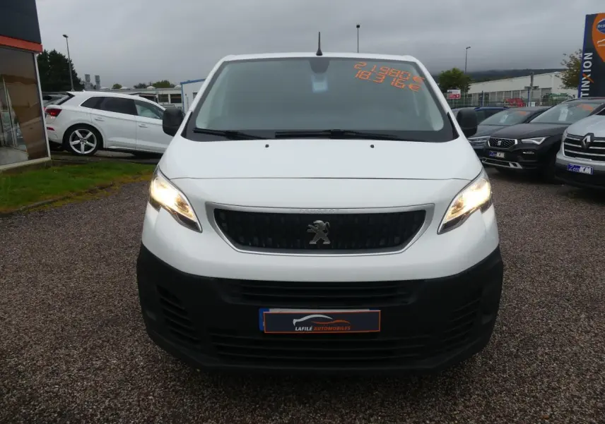 Vue frontale d'un Peugeot Expert Fourgon blanc avec calandre noire et phares allumés sur parking.