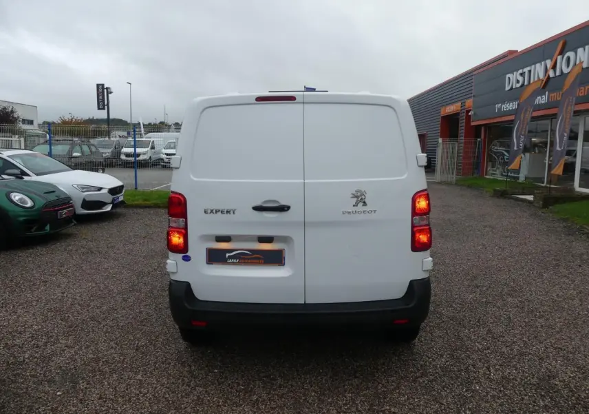 Vue arrière d'un Peugeot Expert Fourgon blanc avec feux allumés sur parking devant un bâtiment commercial.
