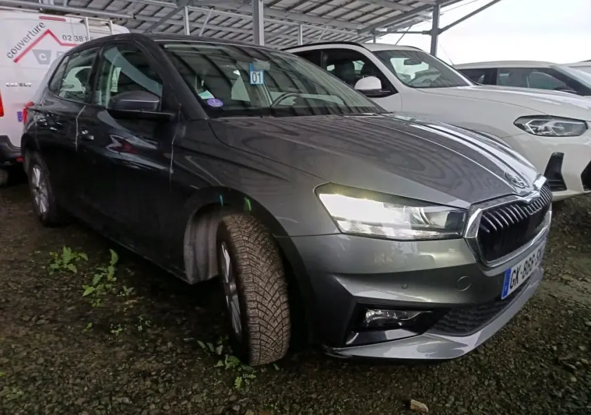 Vue 3/4 avant gauche d'une Skoda Fabia gris graphite 2022 avec phares allumés, stationnée sur sol gravillonné.