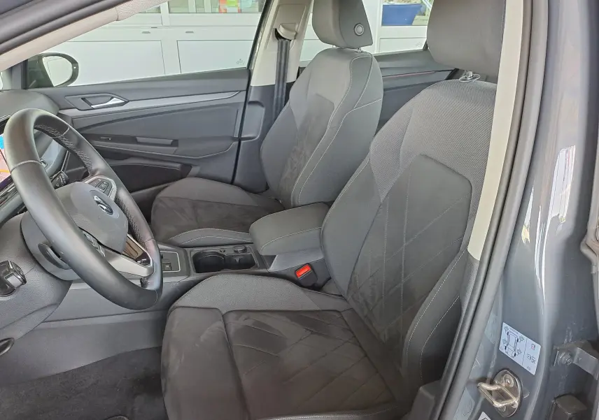 Vue intérieure côté conducteur de la Volkswagen Golf VIII gris dauphin, sièges en tissu noir avec surpiqûres blanches.