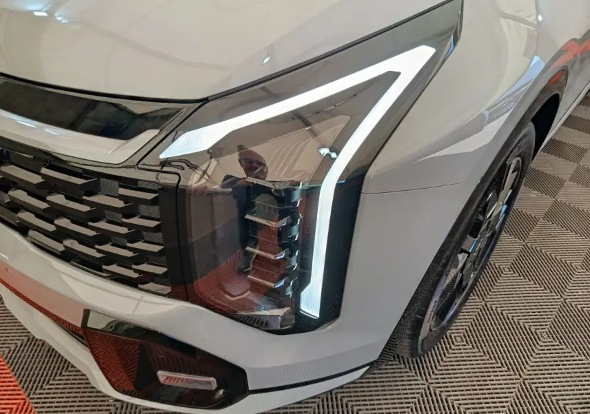 Gros plan sur le phare avant droit allumé du KIA Sportage gris clair, avec calandre noire et jante visible.