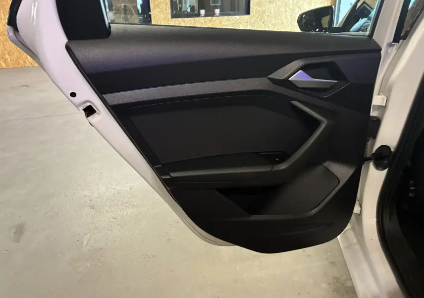 Vue rapprochée de la porte arrière gauche noire ouverte d’un Audi A1 blanc, avec poignée intérieure argentée.