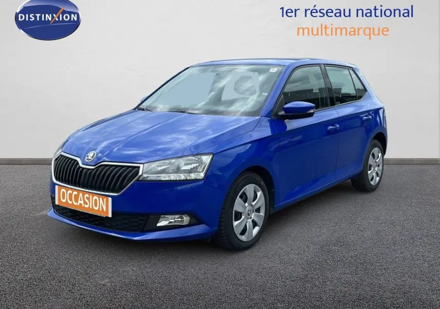 Skoda Fabia 1.0 TSI bleu énergie vue 3/4 avant, garée devant un garage avec façade vitrée et enseignes rouges.