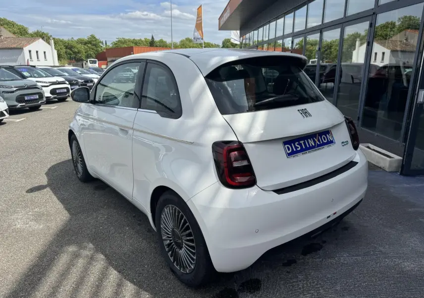 Vue 3/4 arrière droite d'une Fiat 500 électrique blanche avec jantes alliage et toit panoramique, stationnée devant un showroom.