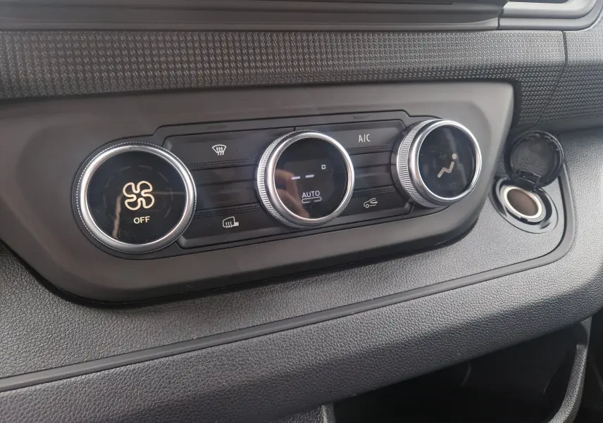 Détail de la console de climatisation du Renault Trafic Fourgon gris urbain, avec commandes rotatives et prise allume-cigare ouverte.
