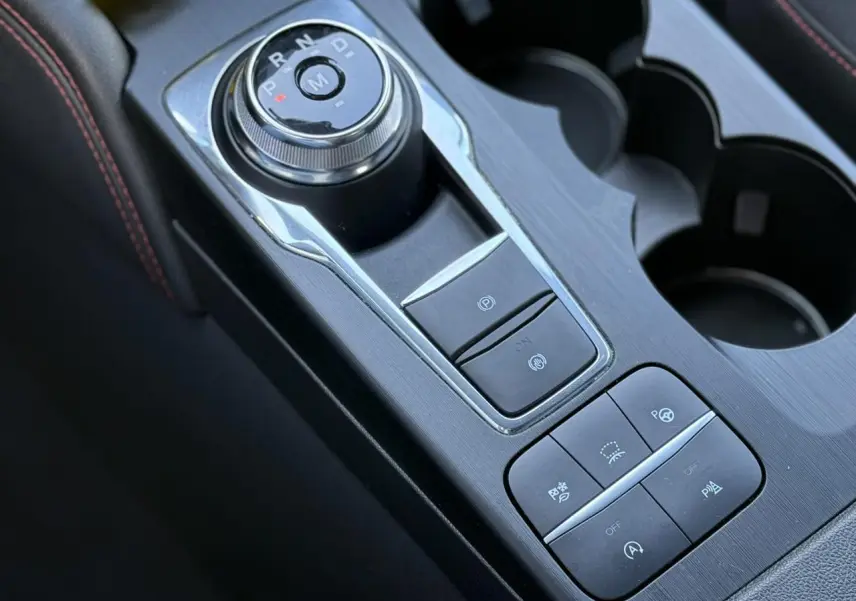 Gros plan sur la console centrale du Ford Kuga 2024 rouge Lucifer, avec sélecteur rotatif de boîte auto et commandes d’assistance.