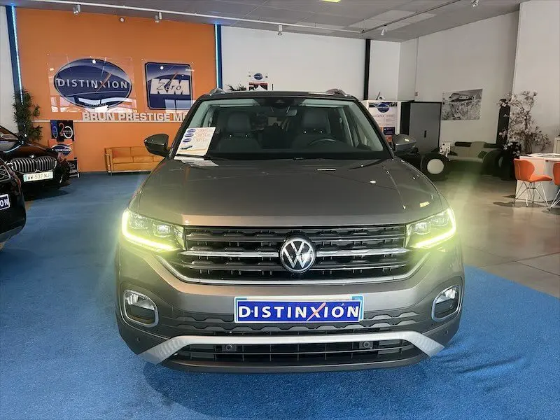 Vue frontale d'un Volkswagen T-Cross gris Limestone métallisé avec phares allumés en showroom Distinxion.