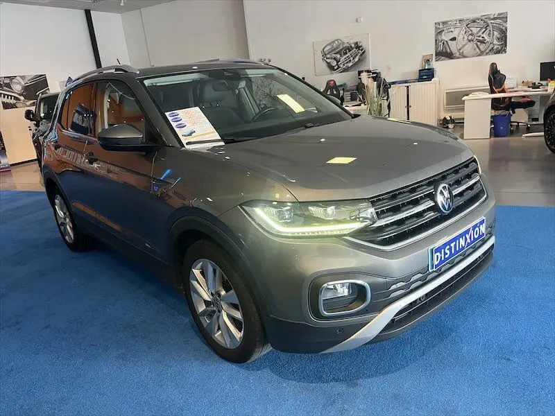 Volkswagen T-Cross gris Limestone métallisé vu en 3/4 avant droit dans un showroom avec phares allumés.