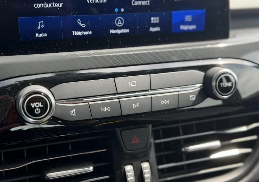 Gros plan sur la console centrale du Ford Kuga 2024, boutons de contrôle audio et écran tactile visible en arrière-plan.
