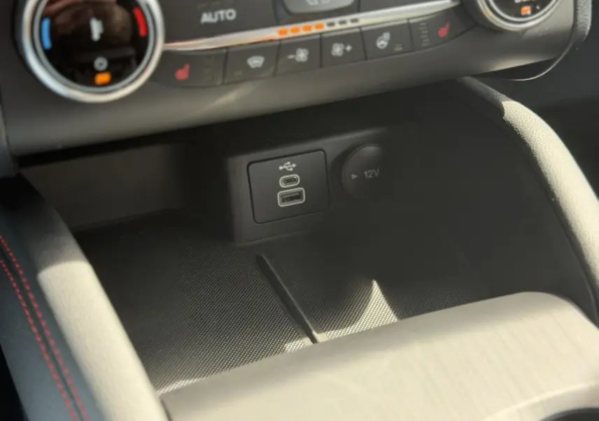 Gros plan sur la console centrale du Ford Kuga ST-Line 2024, montrant ports USB et prise 12V avec surpiqûres rouges.