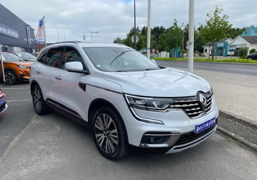 Renault Koleos blanc en 3/4 avant droit, avec calandre chromée et jantes alliage distinctives, stationné en extérieur.