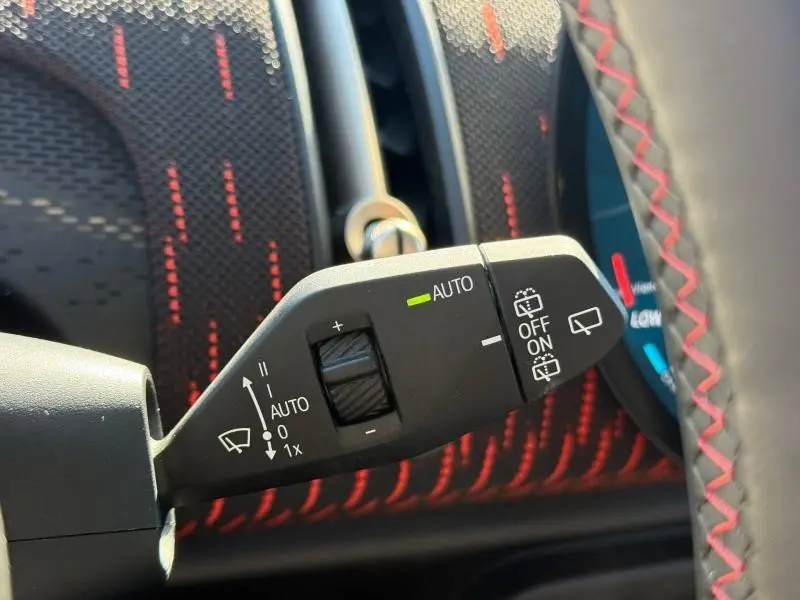 Gros plan sur la commande d'essuie-glace avec réglage automatique, volant noir avec surpiqûres rouges en arrière-plan.
