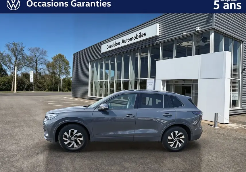 Vue de profil côté gauche d’un Volkswagen Tiguan gris 2025 garé devant un bâtiment moderne avec grandes baies vitrées.