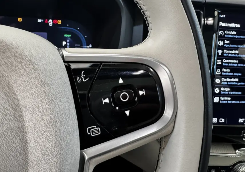 Gros plan sur les commandes du volant cuir beige du Volvo XC60 bleu, affichage numérique et écran tactile visibles.