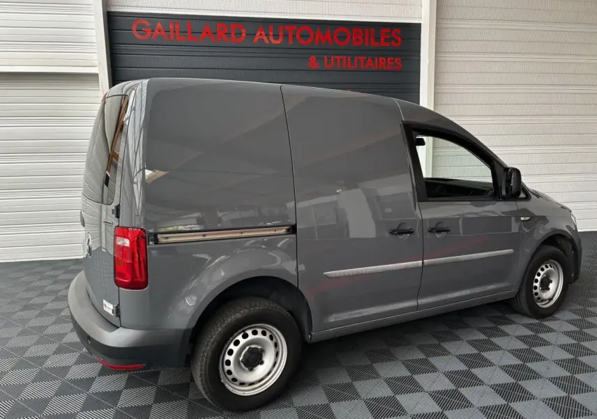 Vue 3/4 arrière côté gauche d'un utilitaire Volkswagen Caddy gris foncé stationné en intérieur sur sol damier.