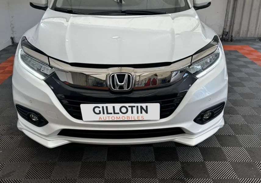 Vue avant rapprochée d’un Honda HR-V blanc avec calandre noire brillante et feux LED, sur sol damier gris et orange.