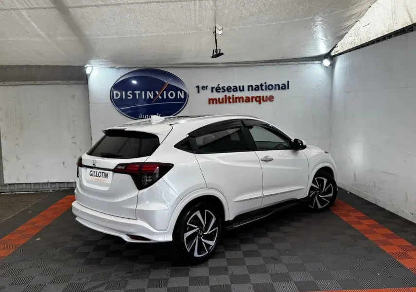 Honda HR-V blanc en vue 3/4 arrière droit, avec jantes noires et toit spoiler dans un showroom.
