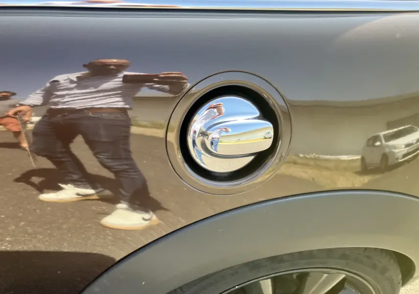 Gros plan sur la trappe à carburant chromée d'une MINI Cooper S R56 marron, reflétant l'environnement extérieur.