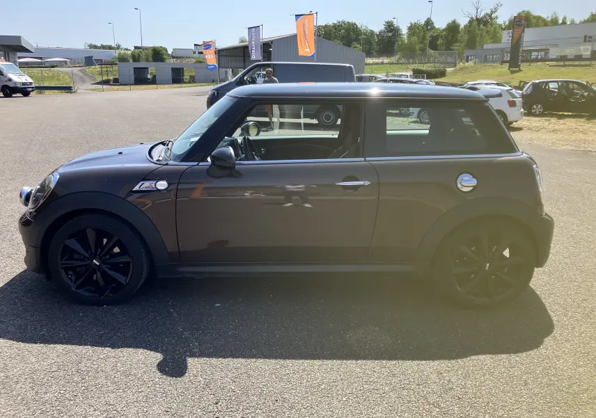 Profil côté gauche d'une MINI Cooper S R56 marron de 2012 avec jantes noires et toit noir sur parking extérieur.