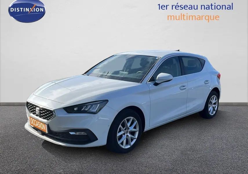 SEAT Leon blanc neige vue 3/4 avant droit, avec jantes alliage et calandre distinctive, stationnée sur sol gris.