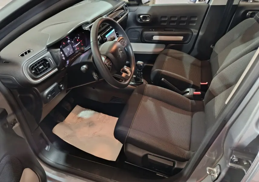 Vue intérieure côté conducteur d'une Citroën C3 gris clair 2024, montrant le tableau de bord et les sièges tissu noirs.
