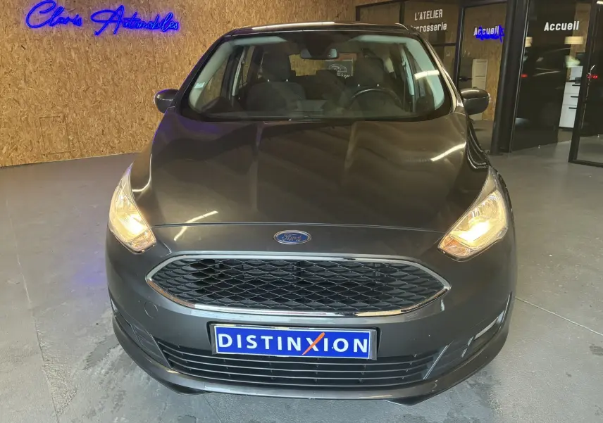 Vue de face d'une Ford C-MAX gris foncé avec phares allumés, stationnée en intérieur devant un mur en bois.