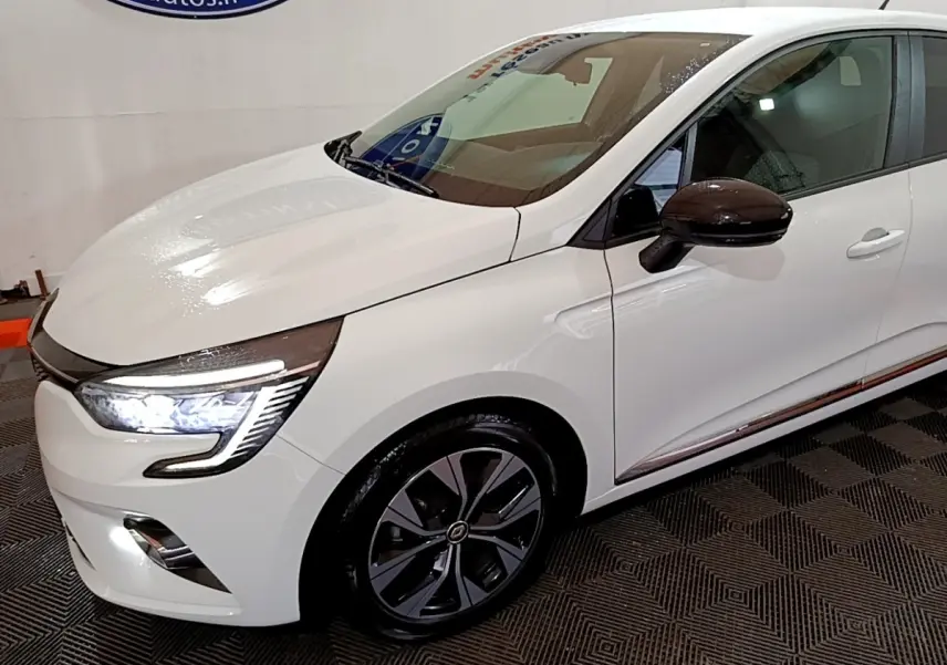 Vue 3/4 avant droit d'une Renault Clio Business blanche avec phares LED allumés et rétroviseurs noirs brillants.