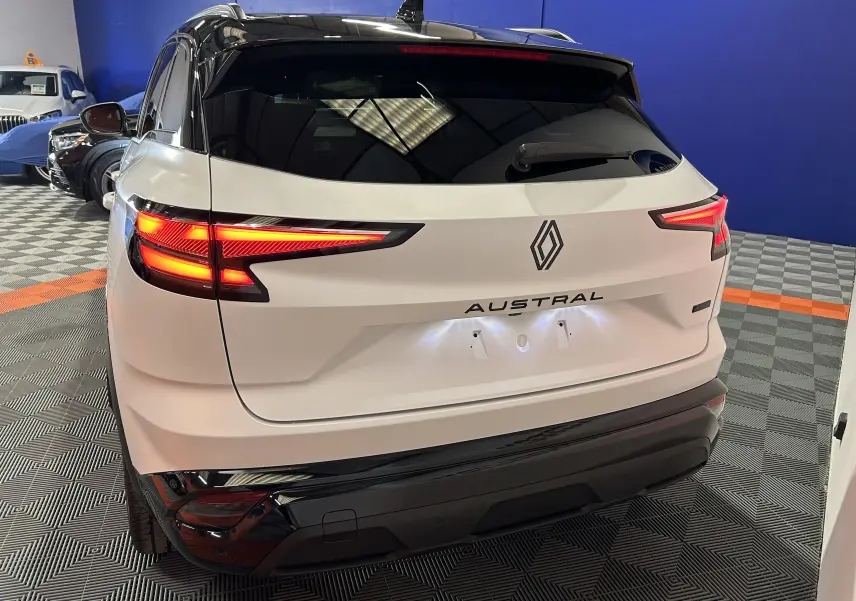 Vue 3/4 arrière d'un Renault Austral blanc nacré satin avec feux arrière LED allumés dans un showroom.