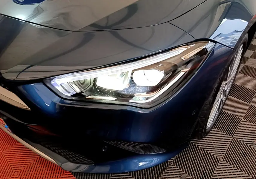 Gros plan sur le phare avant droit allumé d'une Mercedes CLA 180 d Business Line bleu foncé, avec jante visible.