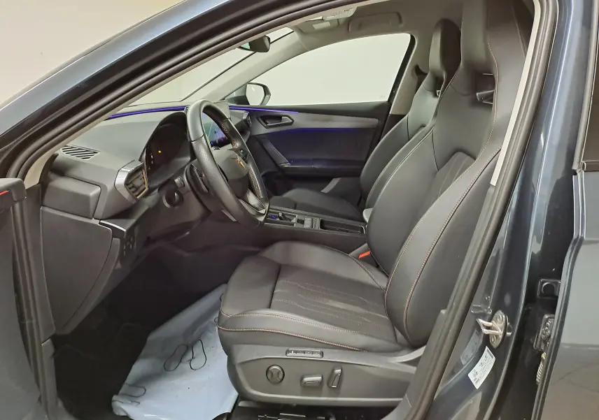 Vue intérieure côté conducteur de la CUPRA Leon 1.5 TSI 150 DS7 V 2023 en gris magnétique, sièges sport noirs avec surpiqûres.