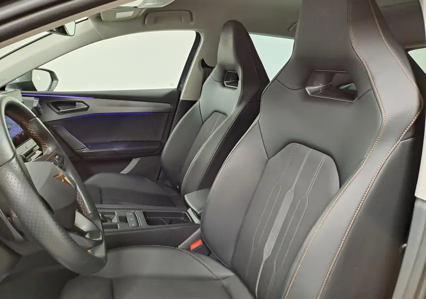 Vue intérieure côté conducteur de la CUPRA Leon 1.5 TSI 150 DS7, sièges sport noirs avec surpiqûres orange et éclairage d'ambiance violet.