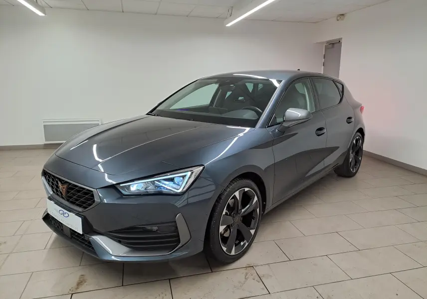 Vue 3/4 avant droite d'une CUPRA Leon 2023 gris magnétique avec jantes noires et phares LED allumés dans un intérieur neutre.