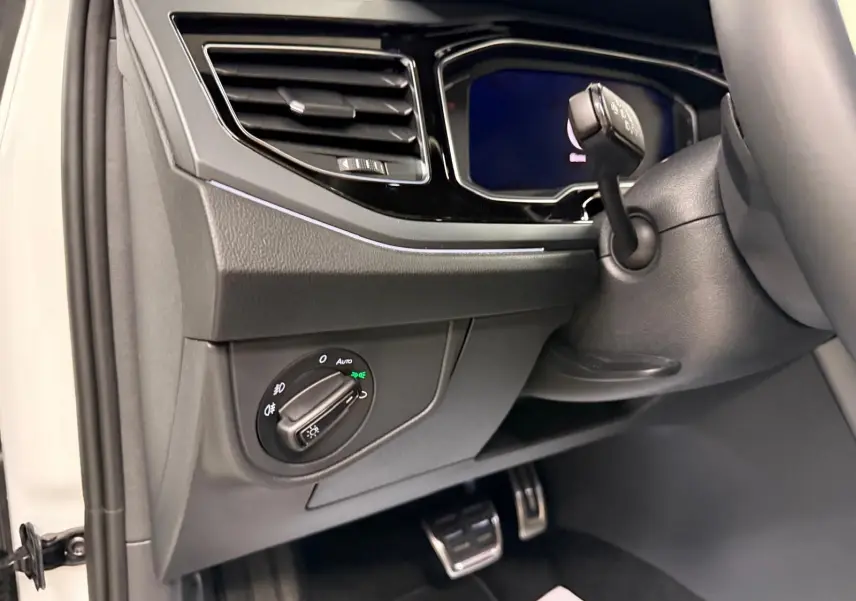 Détail intérieur côté gauche du tableau de bord du Volkswagen Taigo blanc, avec commande des phares et pédalier en acier.