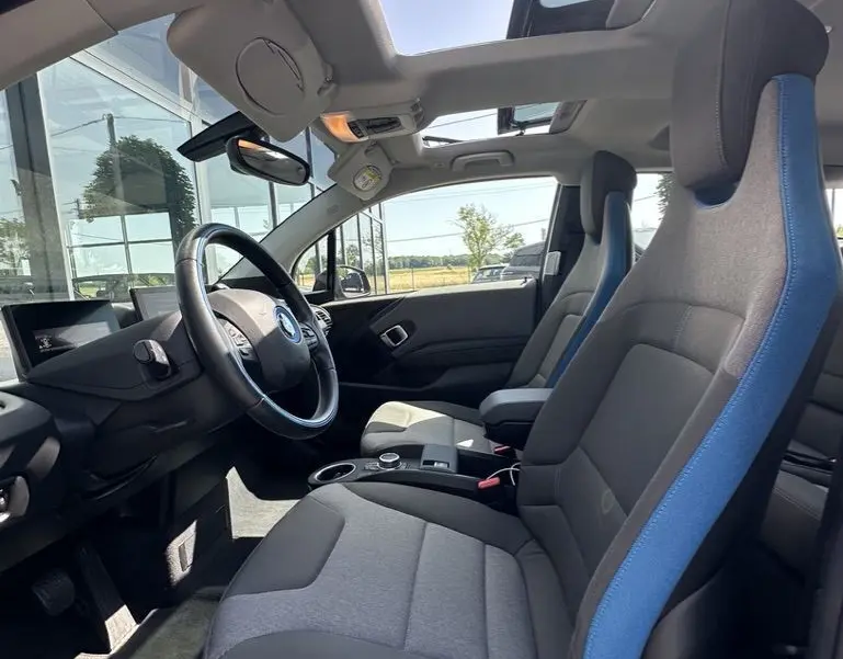 Vue intérieure côté conducteur de la BMW i3 2022 noire, sièges gris avec surpiqûres bleues et toit ouvrant panoramique.