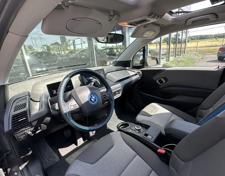 Intérieur de la BMW I3 2022 vue côté conducteur, sièges gris avec surpiqûres bleues et toit ouvrant panoramique.
