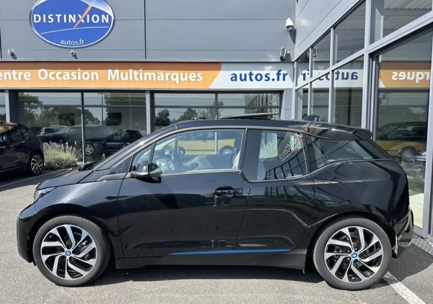 Profil côté gauche d'une BMW i3 noire électrique 2022, avec jantes distinctives et toit vitré, stationnée devant un showroom.