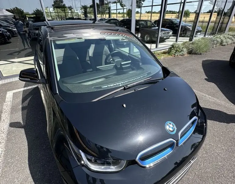 Vue 3/4 avant d'une BMW i3 noire électrique 2022, avec calandre bleue distinctive et pare-brise reflétant un bâtiment vitré.