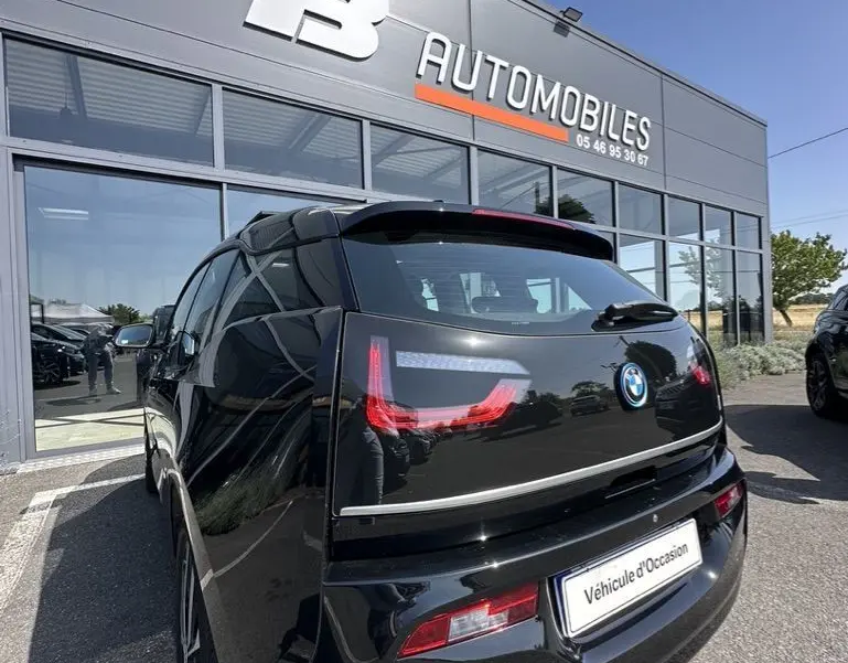 Vue 3/4 arrière d'une BMW I3 noire électrique 2022, avec feux arrière LED distinctifs, stationnée devant un concessionnaire.