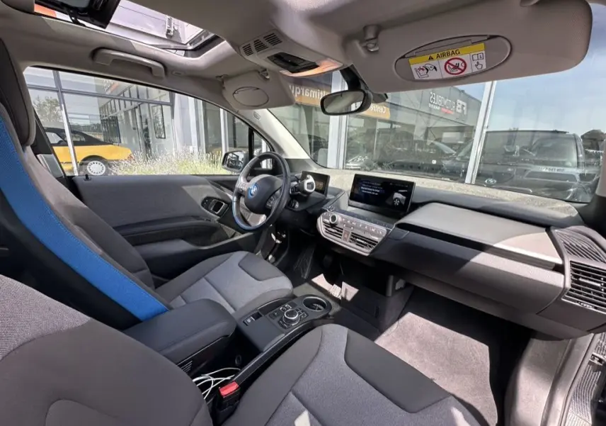 Vue intérieure côté conducteur de la BMW i3 noire 2022, mettant en valeur le tableau de bord moderne et les sièges gris avec appui-tête bleu.