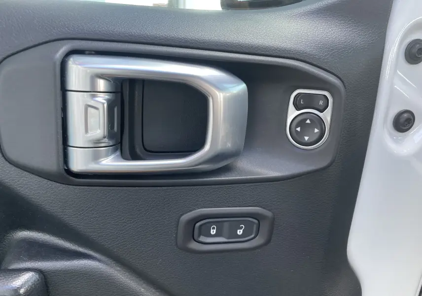 Poignée intérieure argentée et commandes de réglage des rétroviseurs sur la porte noire côté gauche d'un Jeep Wrangler blanc.