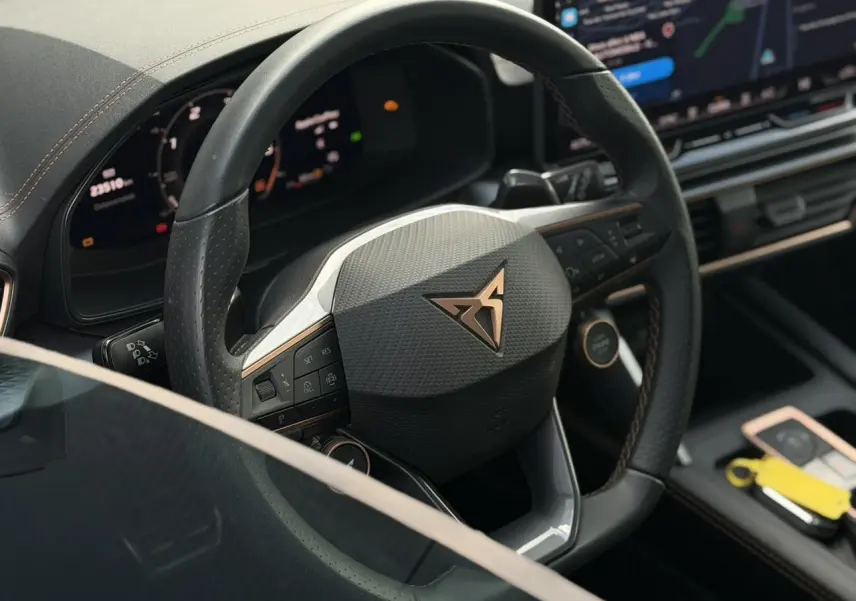 Gros plan sur le volant cuir noir perforé avec logo CUPRA cuivré, tableau de bord digital et console centrale du Formentor blanc.