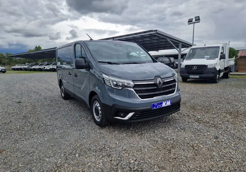 Renault Trafic Fourgon gris urbain vu en 3/4 avant droit sur un parking avec plusieurs véhicules en arrière-plan.