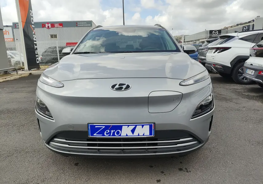 Vue frontale d'un Hyundai Kona électrique gris métallisé avec plaque "Zéro KM" sur un parking extérieur.