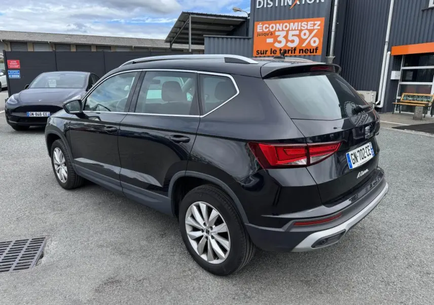 Vue 3/4 arrière droite d'un SUV SEAT Ateca noir métallisé garé devant un bâtiment avec panneaux publicitaires.