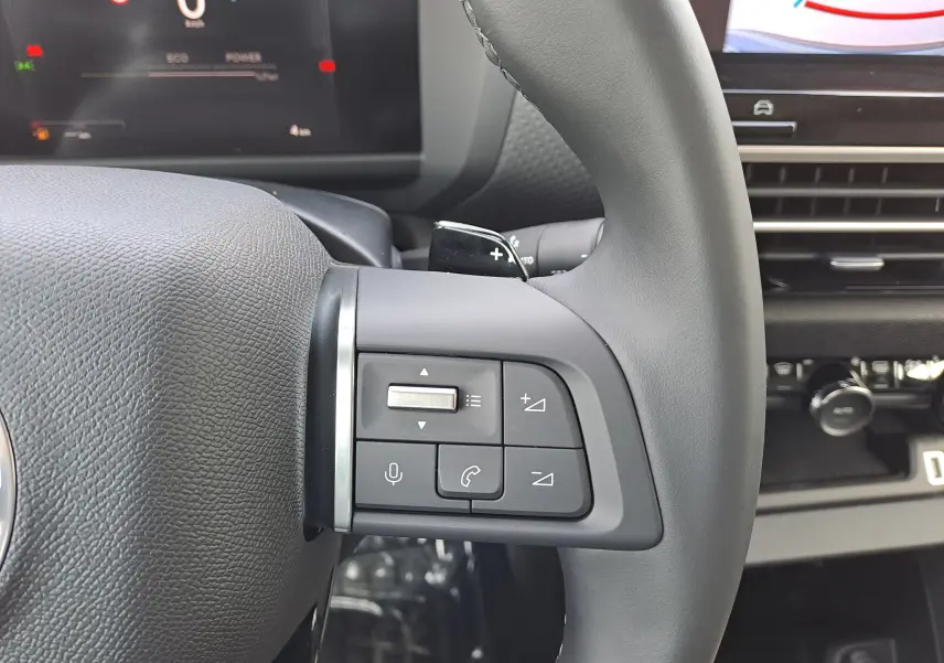 Gros plan sur les commandes au volant côté gauche du Citroën C4 hybride 2025, avec tableau de bord numérique en arrière-plan.