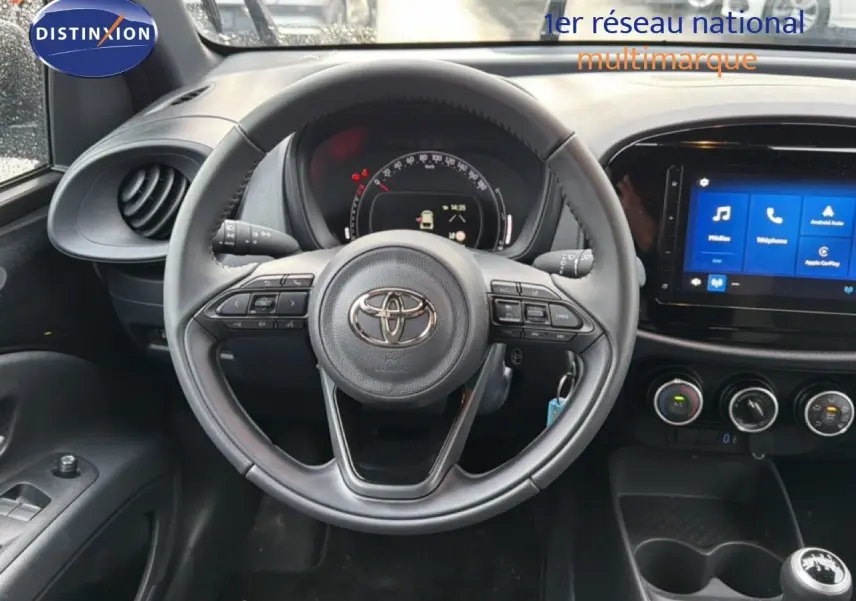 Vue intérieure centrée sur le volant et le tableau de bord noir de la Toyota Aygo X 2025 avec écran tactile et commandes au volant.