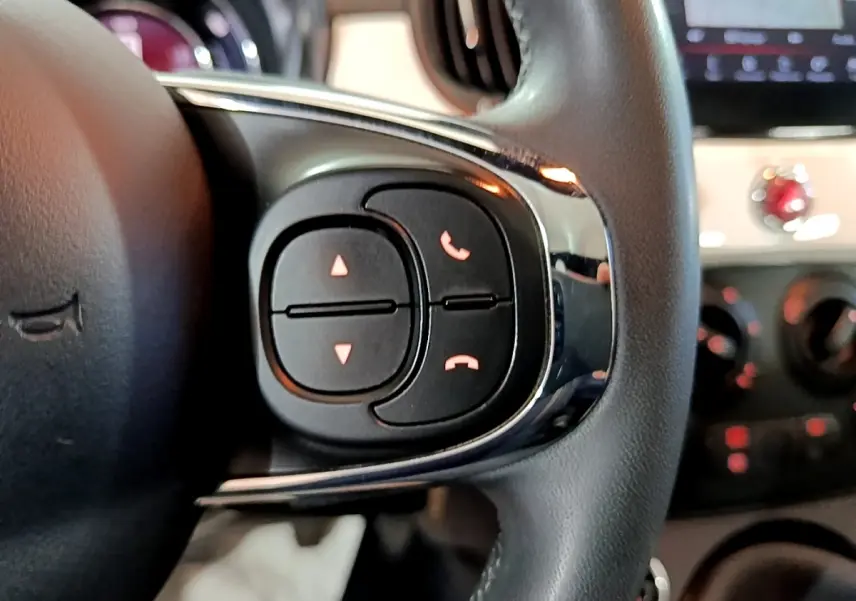 Gros plan sur les commandes du volant cuir noir de la Fiat 500 Hybrid 2021, avec tableau de bord flou en arrière-plan.