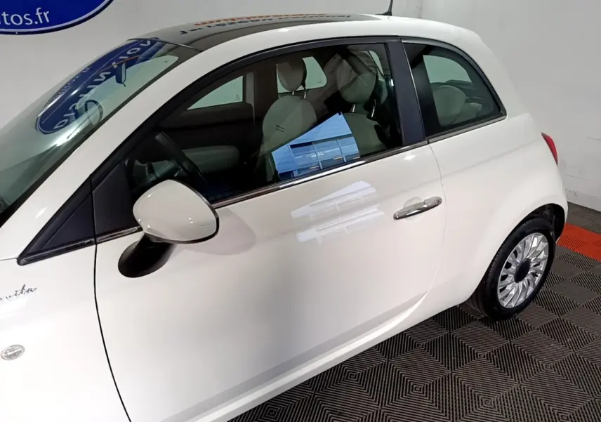 Vue côté gauche d'une Fiat 500 blanche Dolcevita 2021, avec rétroviseur et jante visibles en intérieur showroom.