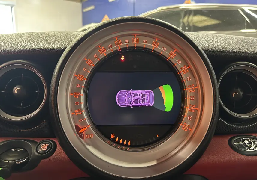 Tableau de bord circulaire de MINI Cabriolet rouge 2012, affichant un écran central avec vue schématique du véhicule et jauge de vitesse orange.